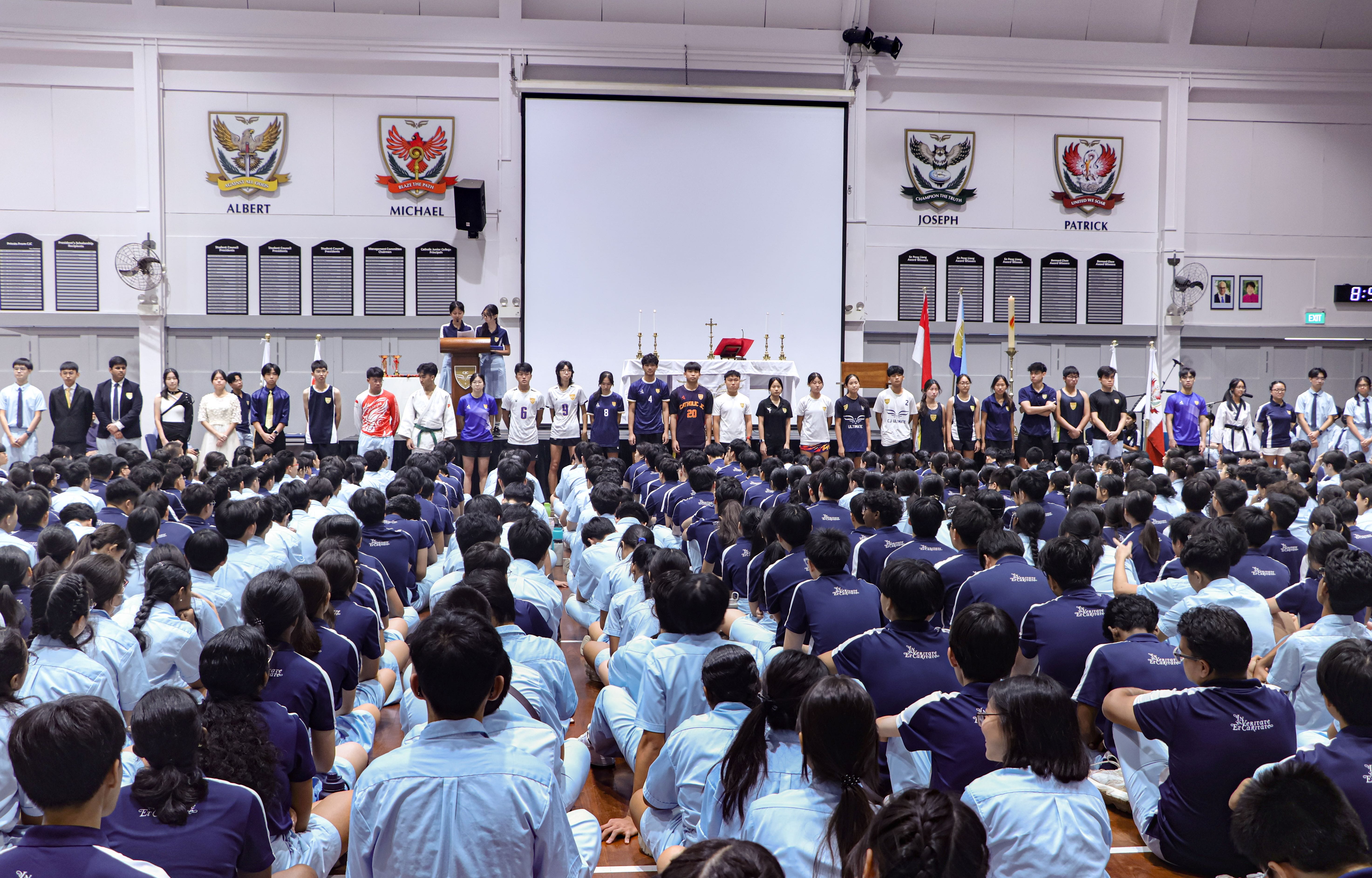 Students in uniforms sit on a hall floor facing a stage with standing students, house crests, and a projector screen.